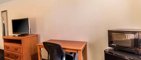 Une petite chambre d’hôtel avec un bureau en bois, une chaise de bureau noire, un téléviseur posé sur une commode en bois et un four à micro-ondes sur un réfrigérateur noir.