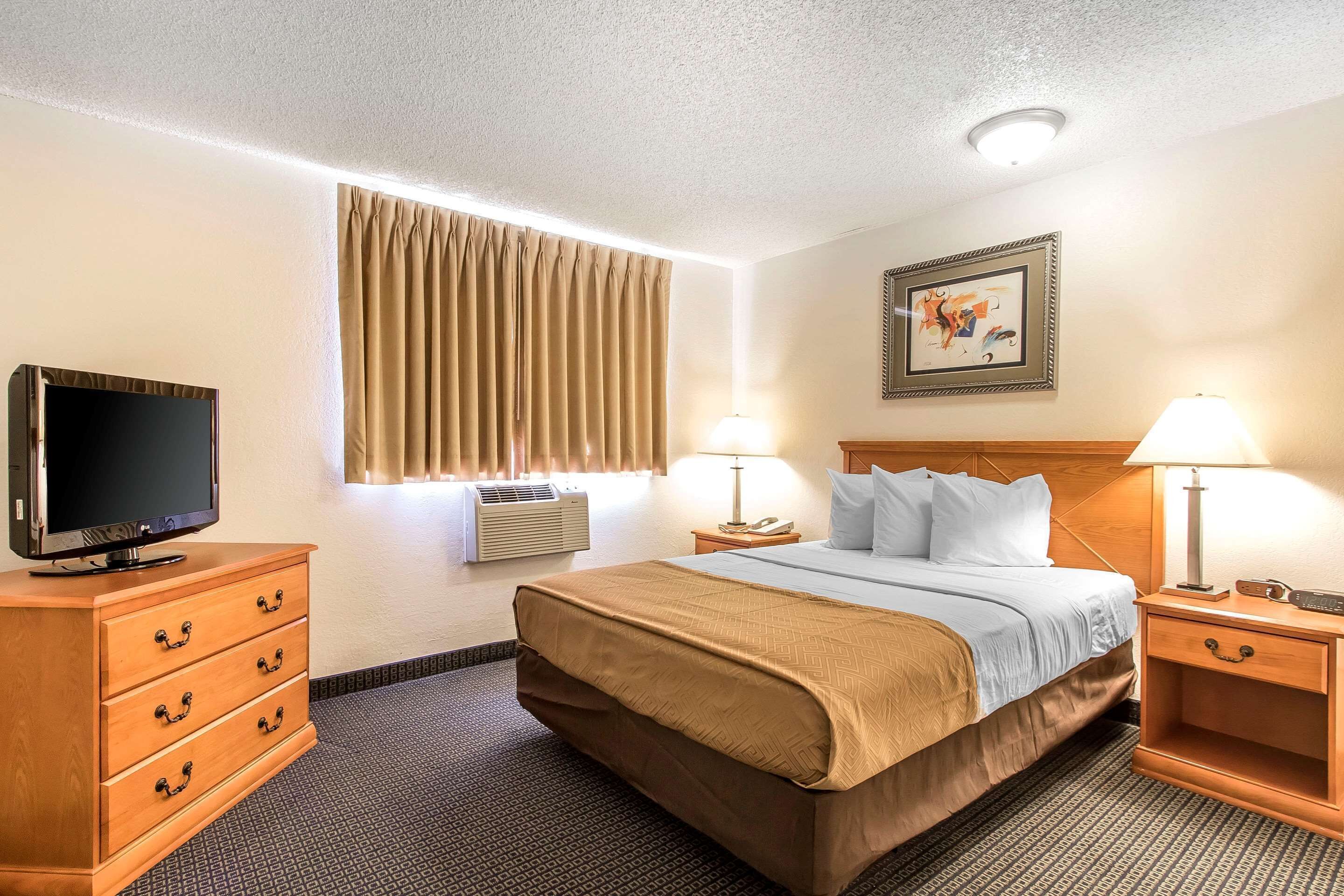 A hotel room with a bed, a TV, a dresser, a nightstand with lamps, an air conditioner, and a framed picture on the wall.