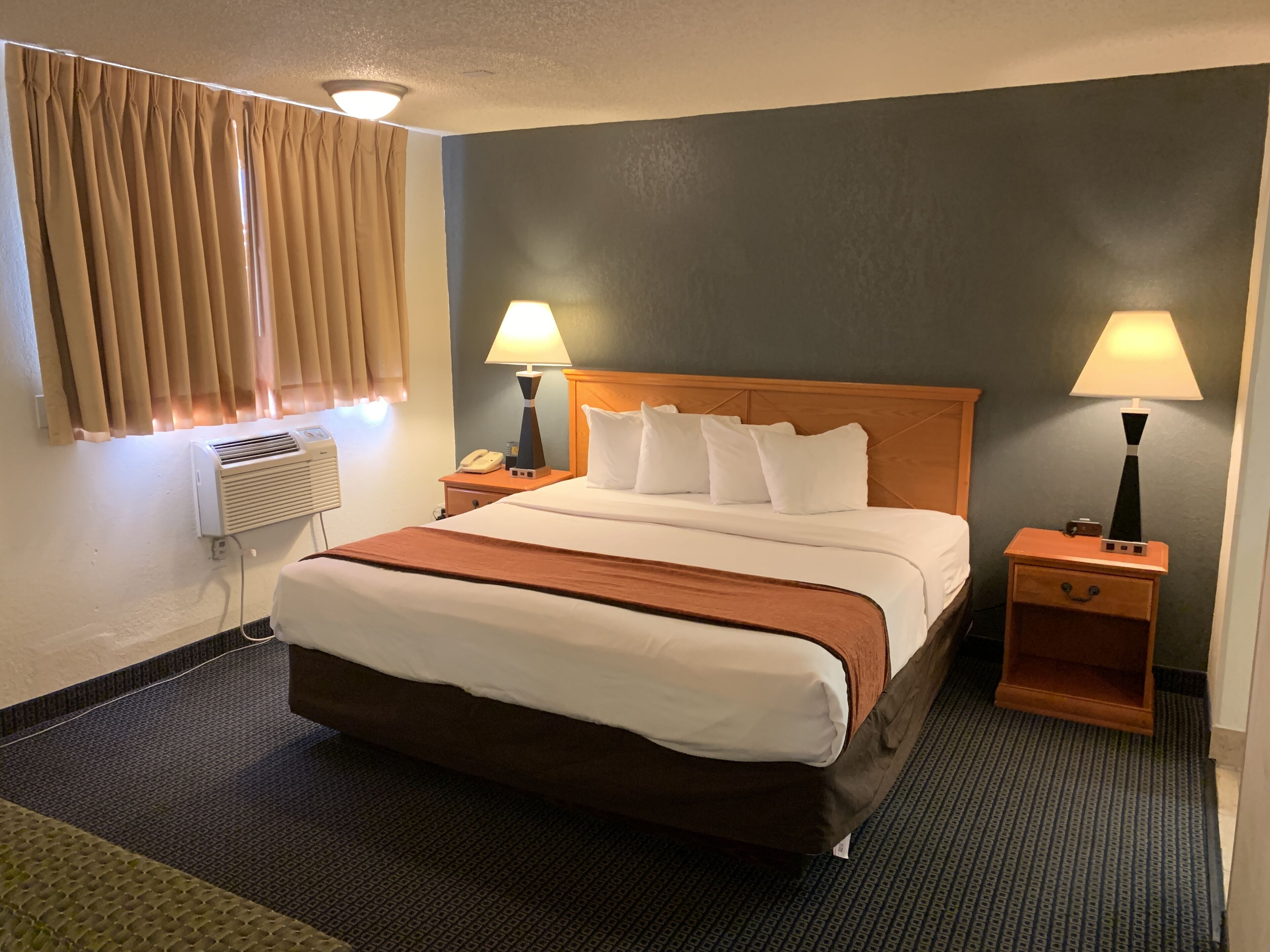 A hotel room with a bed, two bedside tables, a lamp on each table, a wall-mounted heater, and a window with curtains.