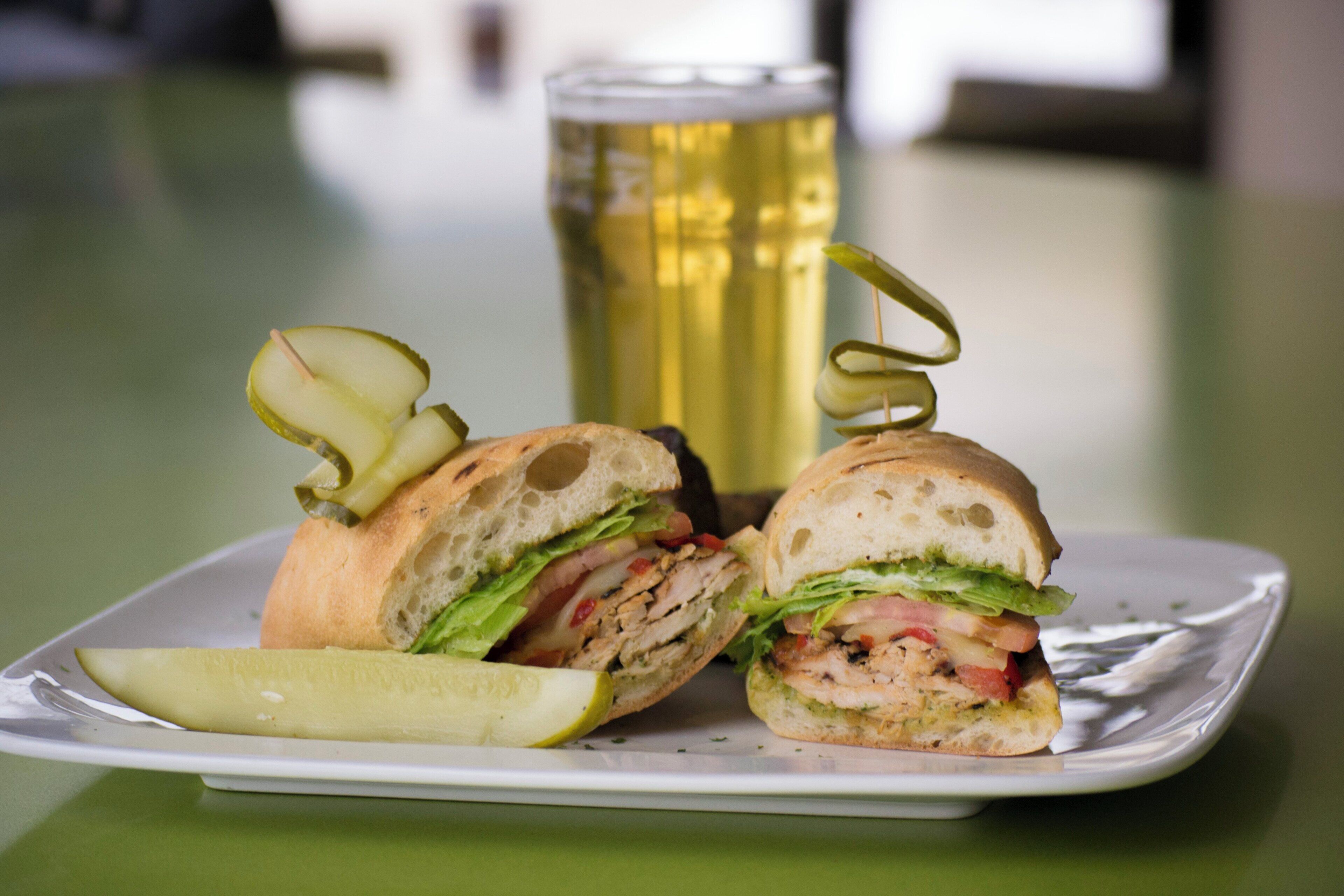 A sandwich cut in half with pickles on a plate, a glass of beer, and a table.