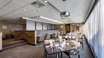 Een restaurantgedeelte in een hotel met meerdere tafels en stoelen, een bar in het midden en een trap naar een hoger niveau.