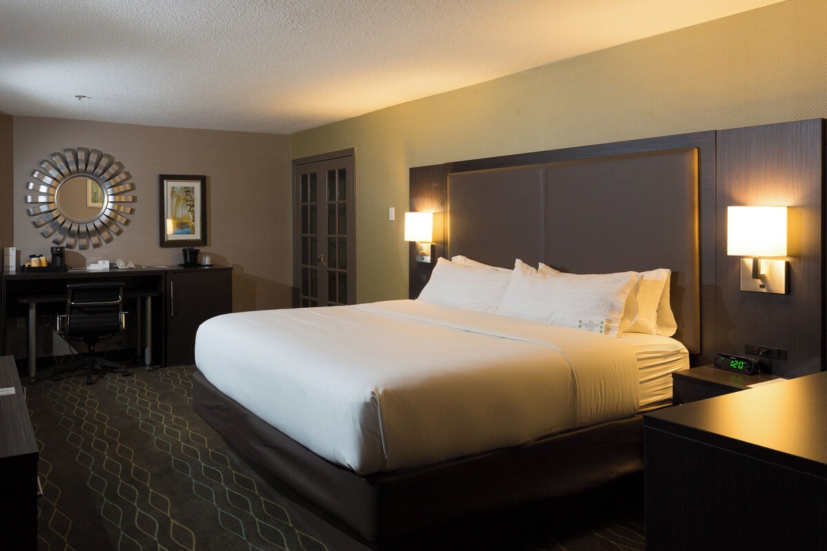 A hotel room with a large bed, a desk, a chair, a round mirror, and two wall-mounted lamps.