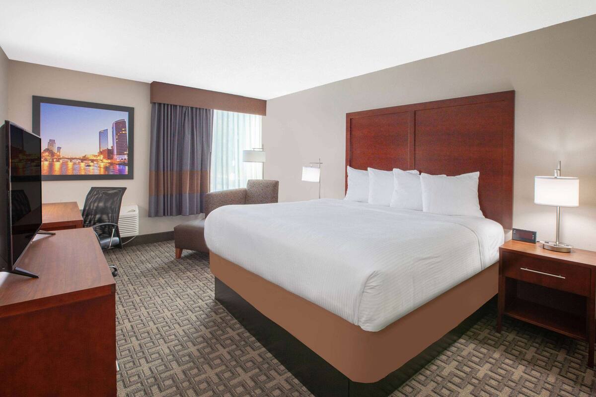 Een hotelkamer met een groot bed, een bureau met een televisie, een stoel, een lamp en een stadsbeeld aan de muur.