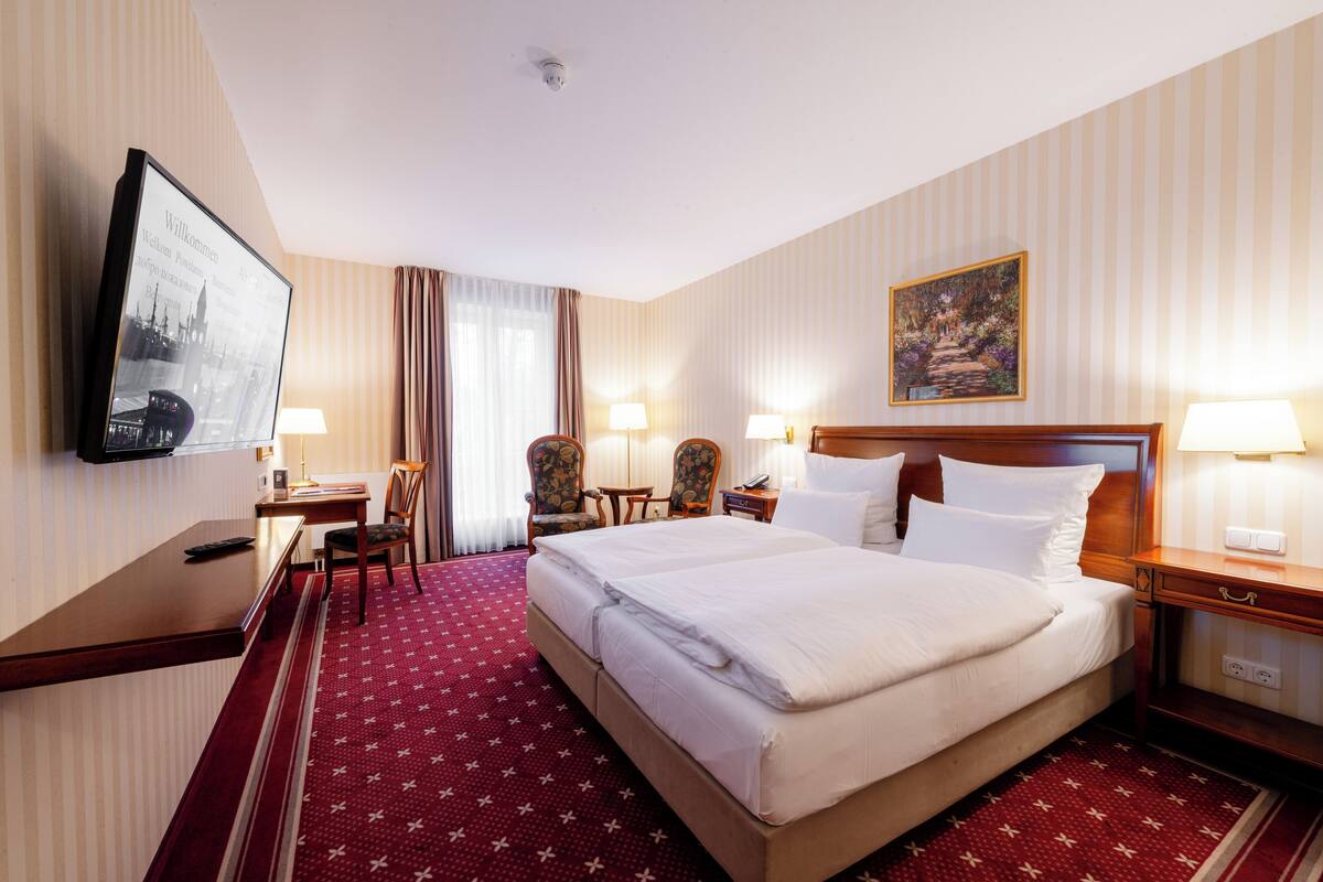 A hotel room with a large bed, two bedside tables with lamps, a desk with a chair, a television mounted on the wall, and a painting on the wall.