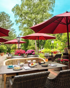 Un área de comedor al aire libre con sombrillas rojas, mobiliario de mimbre y una variedad de platos sobre las mesas.