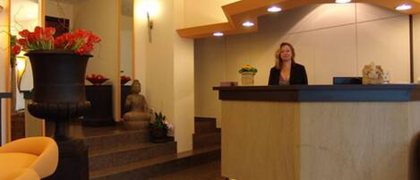 A reception desk with a woman standing behind it, a Buddha statue, and a large potted plant with red flowers.