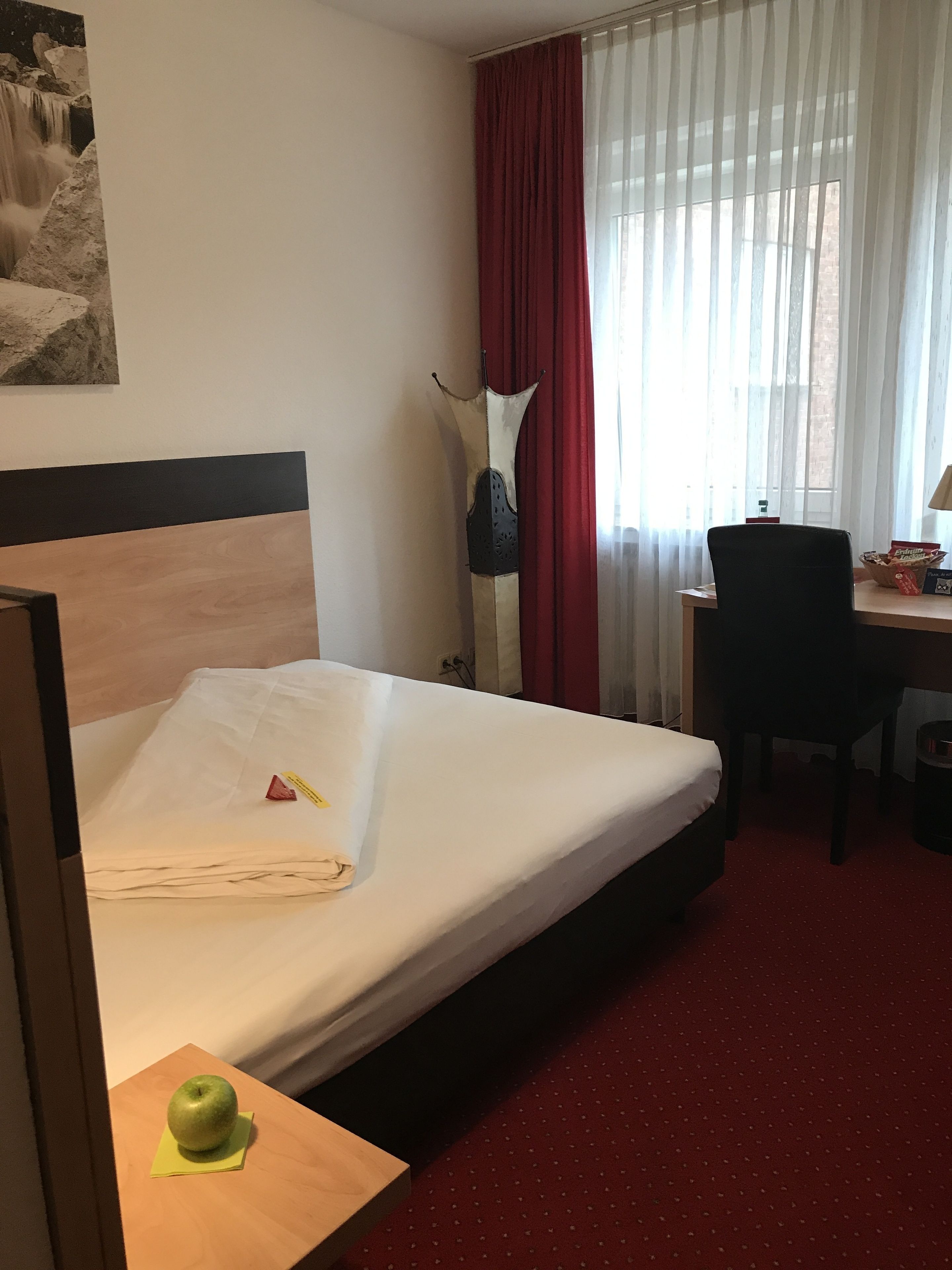 A hotel room with a bed, a desk, a chair, a red carpet, and a window with curtains.