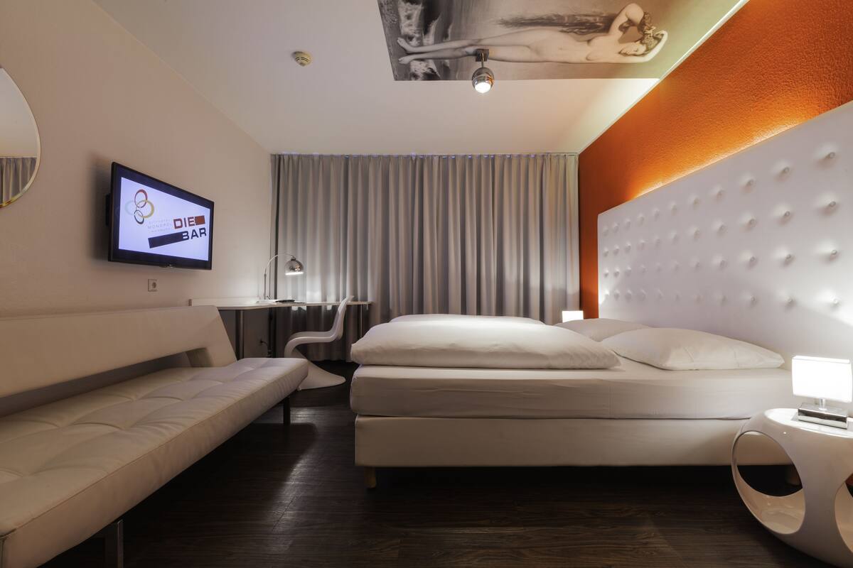 A modern hotel room with a bed, a sofa, a desk, and a TV displaying the hotel's logo.