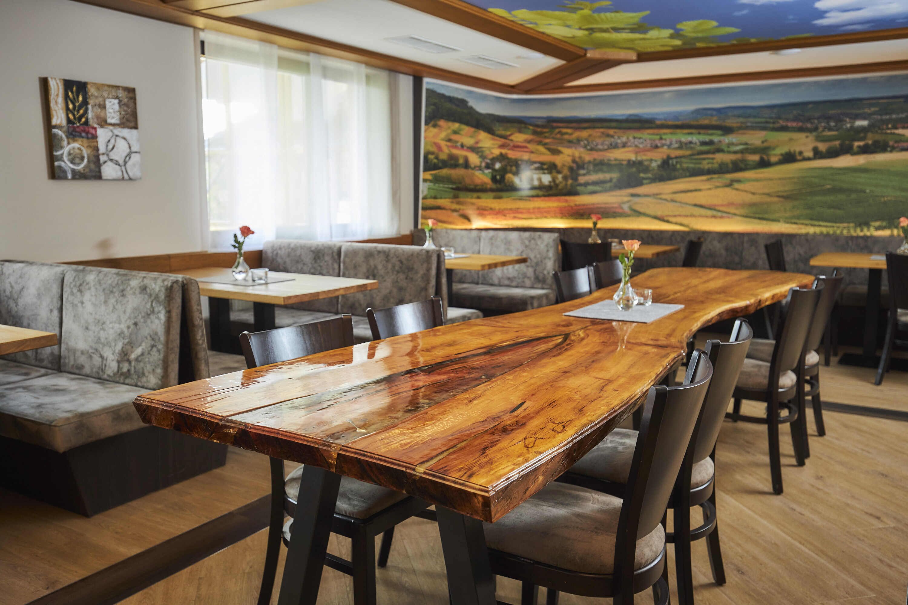Un restaurante con una larga mesa de madera, asientos acolchados y un mural de un paisaje en la pared.