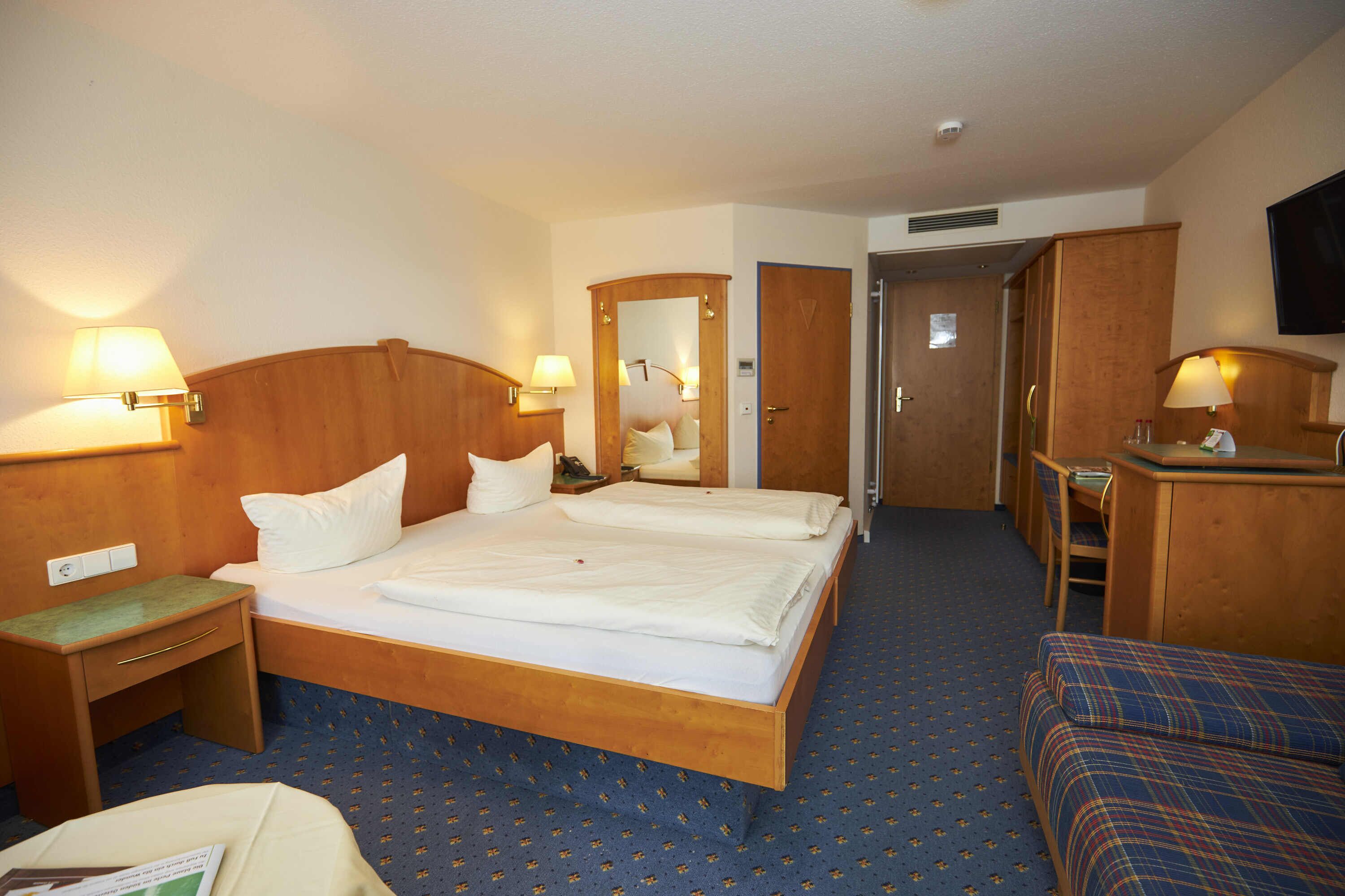 A hotel room with a double bed, a desk with a chair, a small table, and a TV mounted on the wall.
