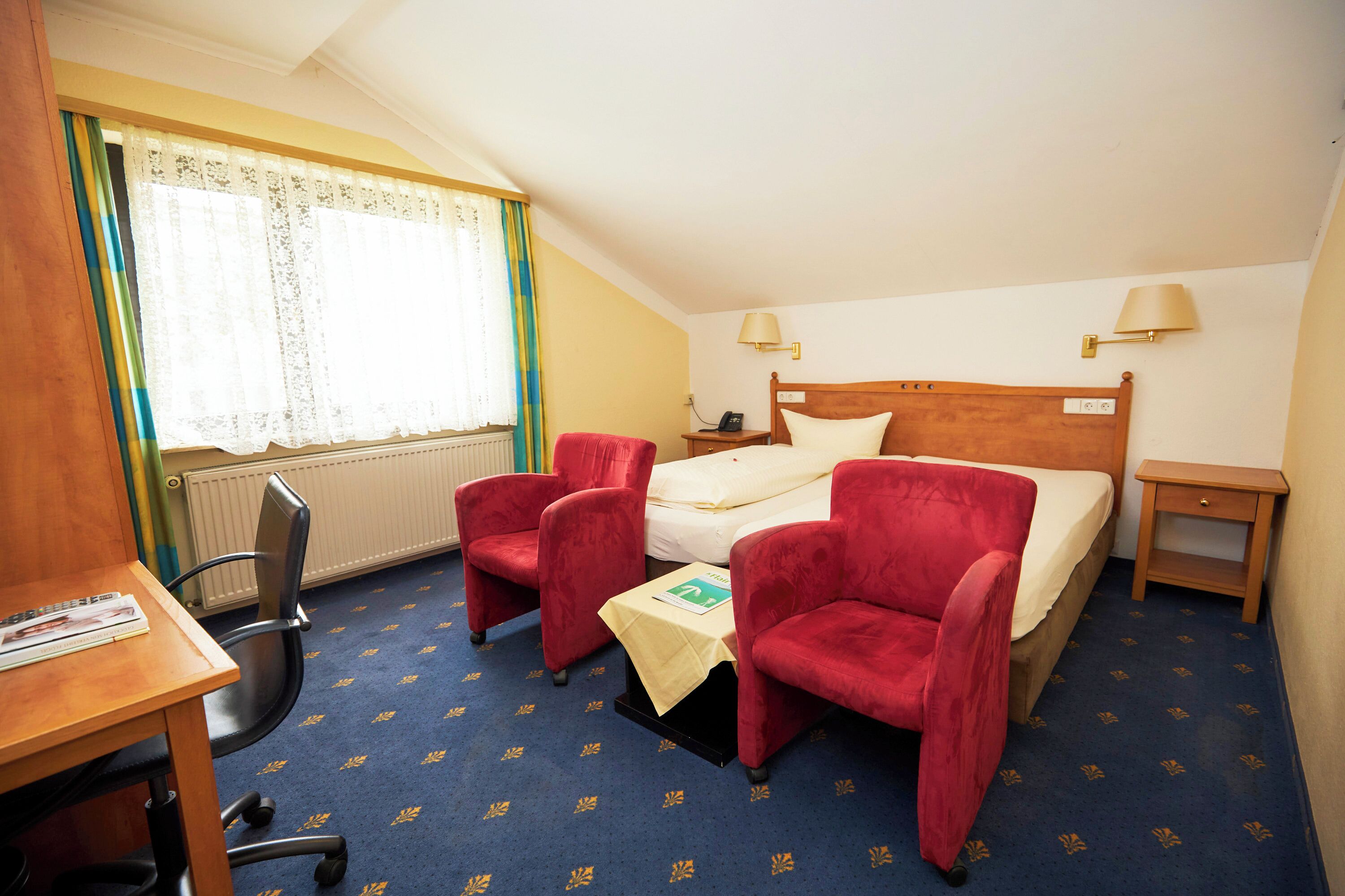 A hotel room with a bed, two red armchairs, a small table with a white tablecloth, a wooden nightstand, and a lamp.