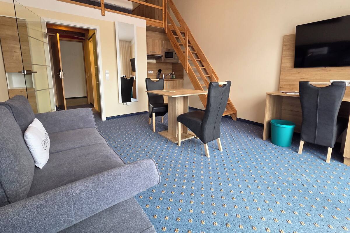 A modern hotel room with a sofa, a dining table, chairs, a TV, a staircase, and a kitchen area.