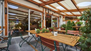 Een terras buiten met houten tafels en stoelen, plantenpotten en een glazen deur naar binnen.