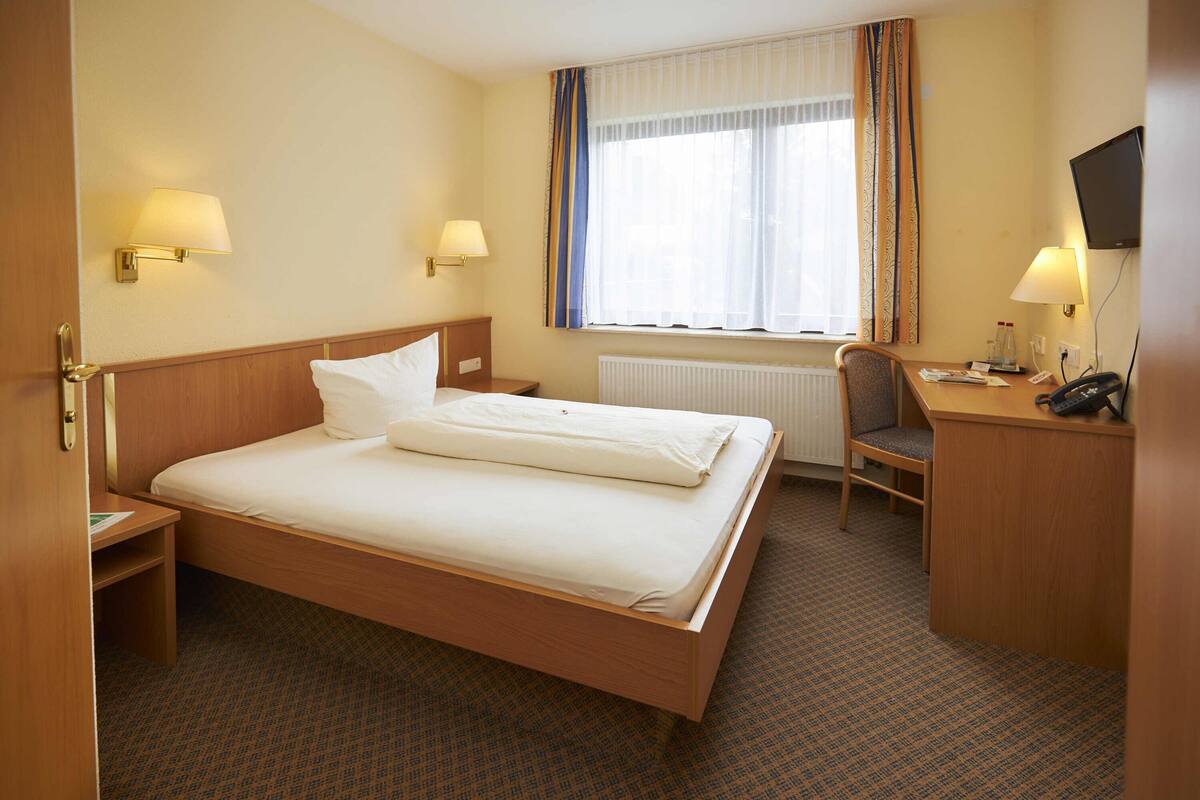 A hotel room with a bed, bedside tables, a desk, a chair, a TV, and a window with curtains.