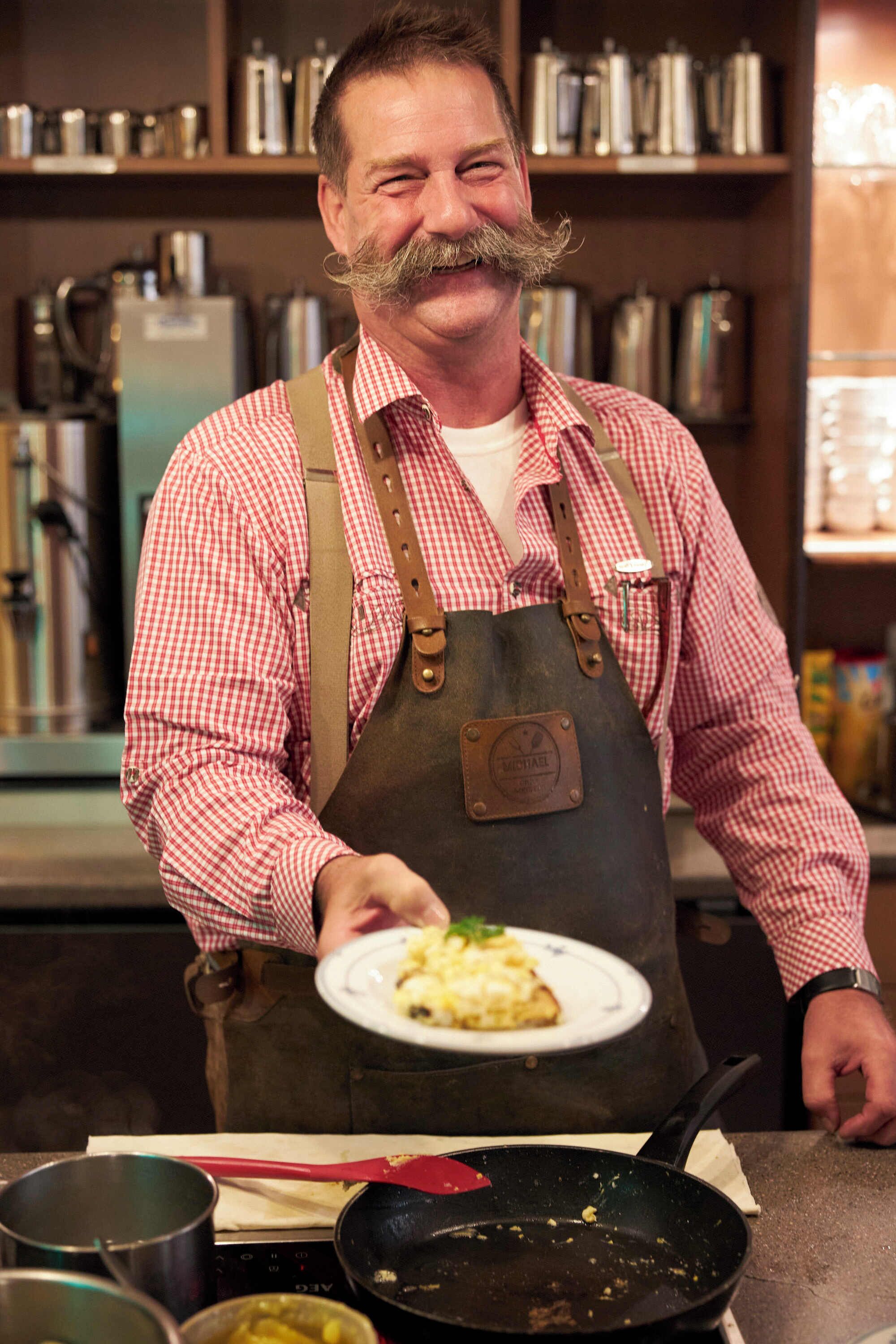 A person in a red and white checkered shirt and apron, holding a plate with food, standing in a kitchen with various cooking utensils and appliances.