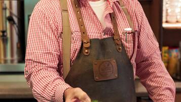 Una persona con camisa y delantal a cuadros rojo y blanco sostiene un plato con comida, de pie en una cocina con diversos utensilios y electrodomésticos.