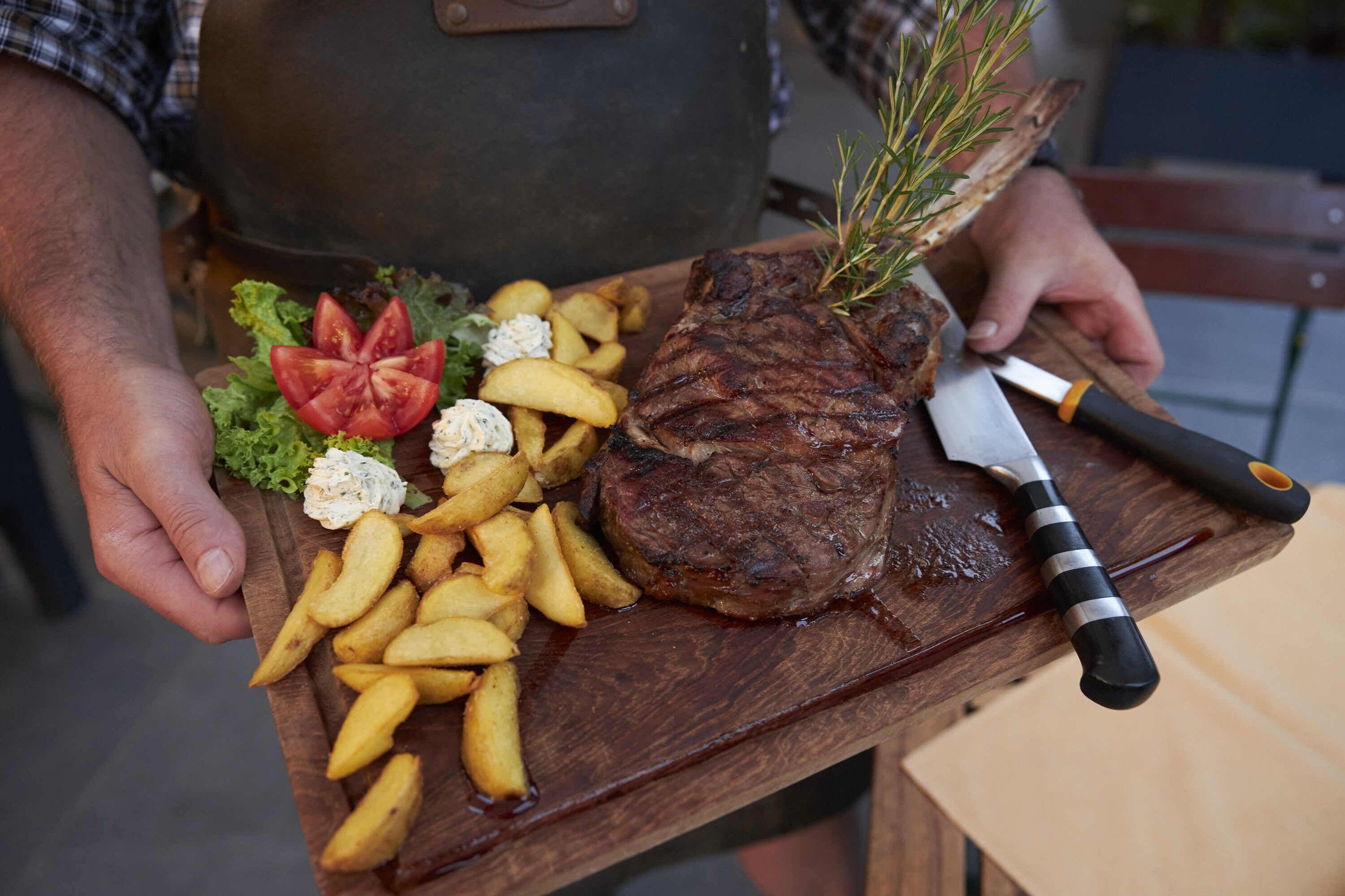 A person holding a wooden board with a steak, fries, and a garnish.