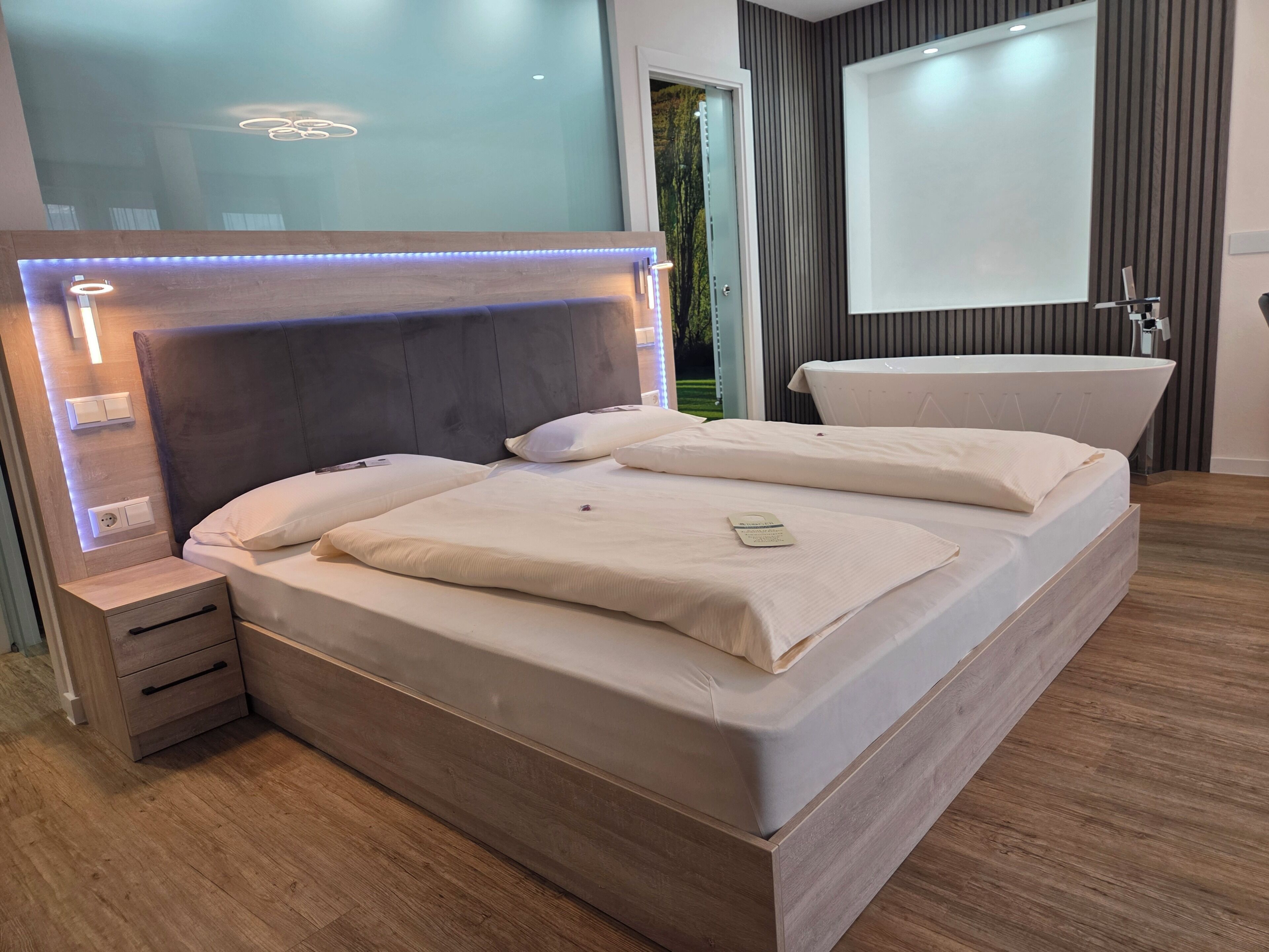 Un dormitorio moderno con una cama grande, mesitas de noche, un banco empotrado, un espejo grande y una bañera.
