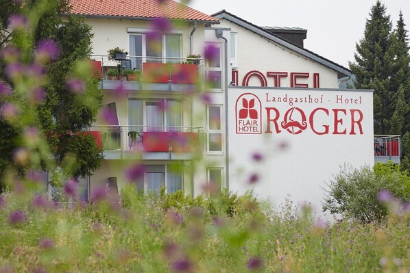 Un hôtel appelé « Flair Hotel Roger », avec une enseigne, des balcons et une végétation luxuriante tout autour.