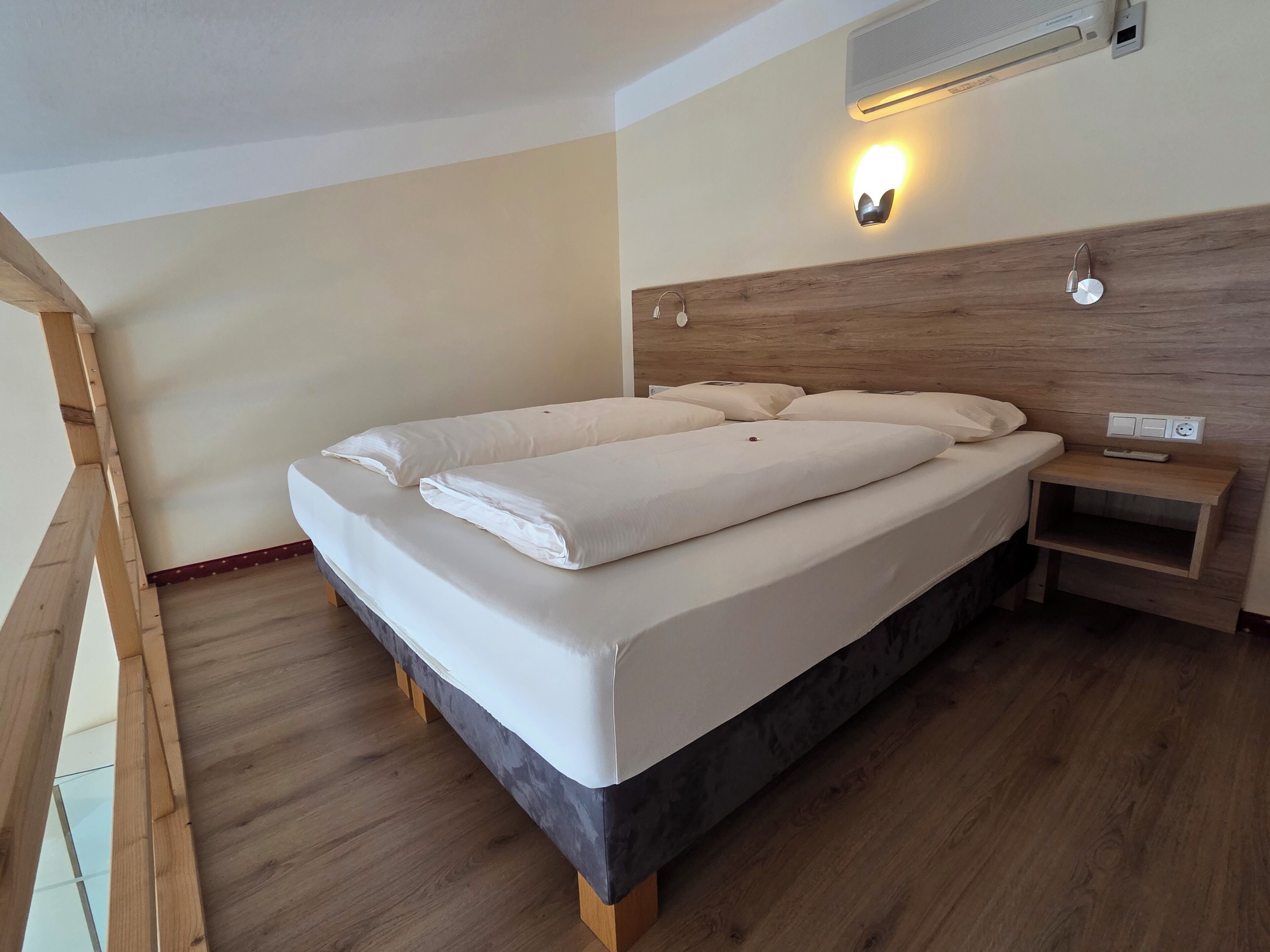 A neatly made bed with white linens, a wooden headboard, a bedside table with a remote control, and a wall-mounted light fixture.