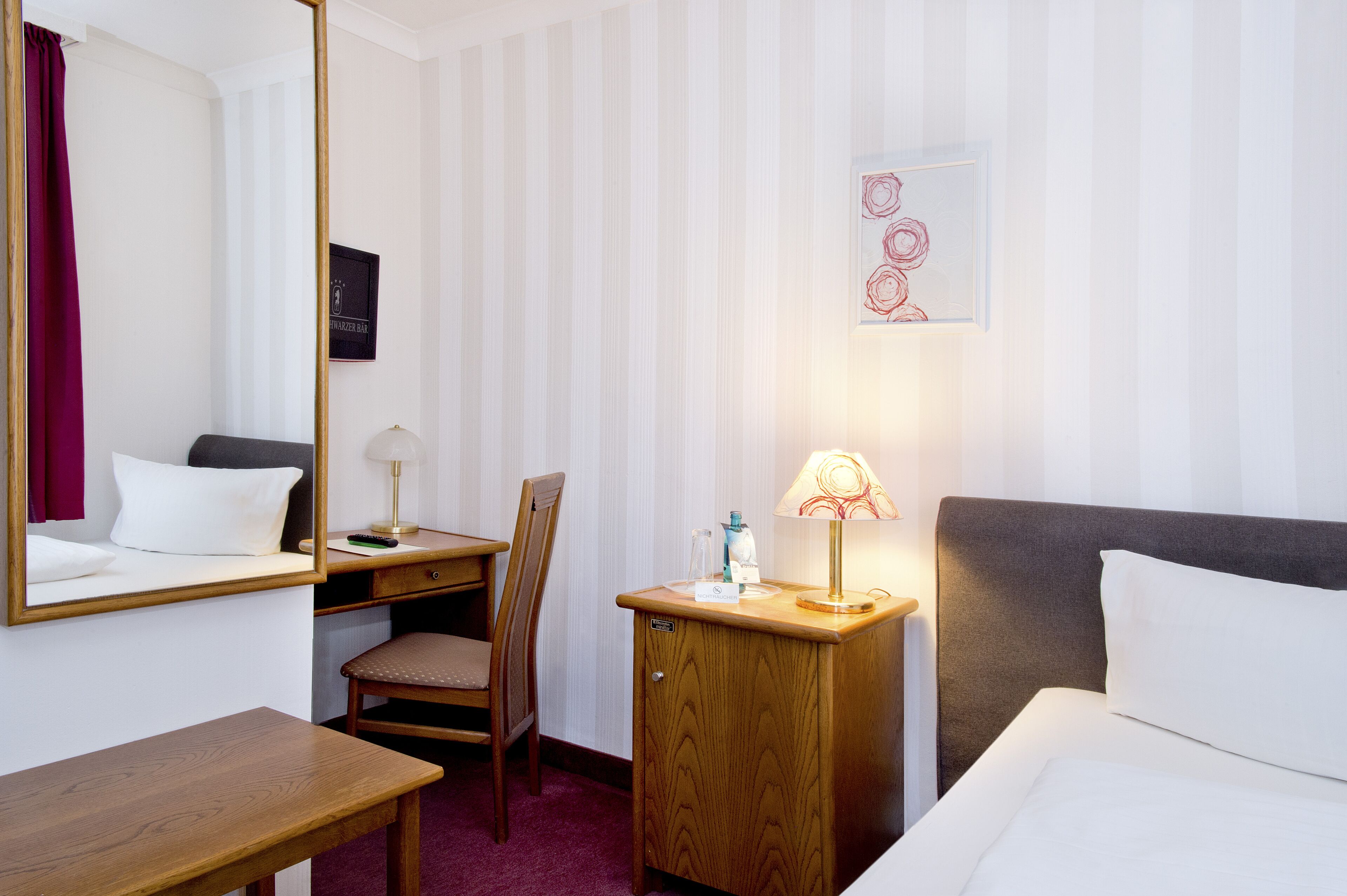 A hotel room with a bed, a desk, a chair, a bedside table, a lamp, and a mirror.