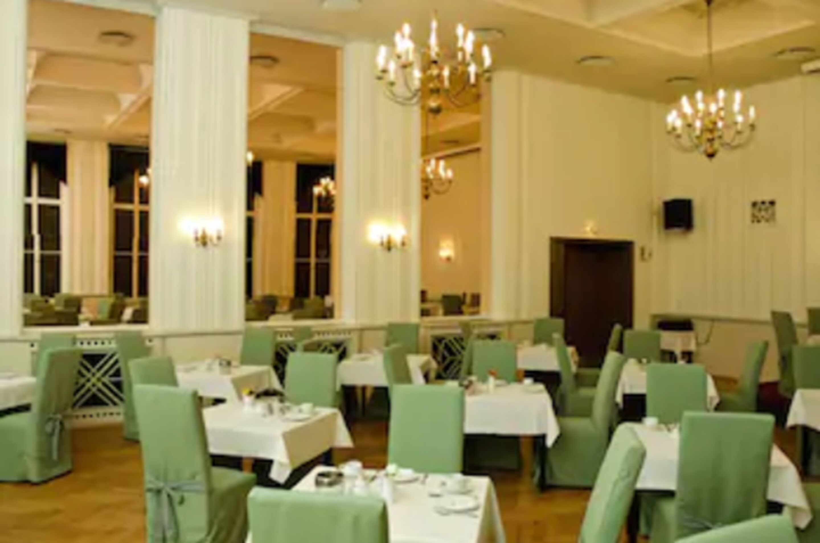 A dining area with green chairs and white tables, large windows, and chandeliers.