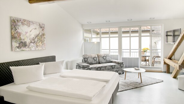 A modern bedroom with two beds, a sofa, a small table, and a large window with a view. - Hotel Schwarzer Bär (Jena)