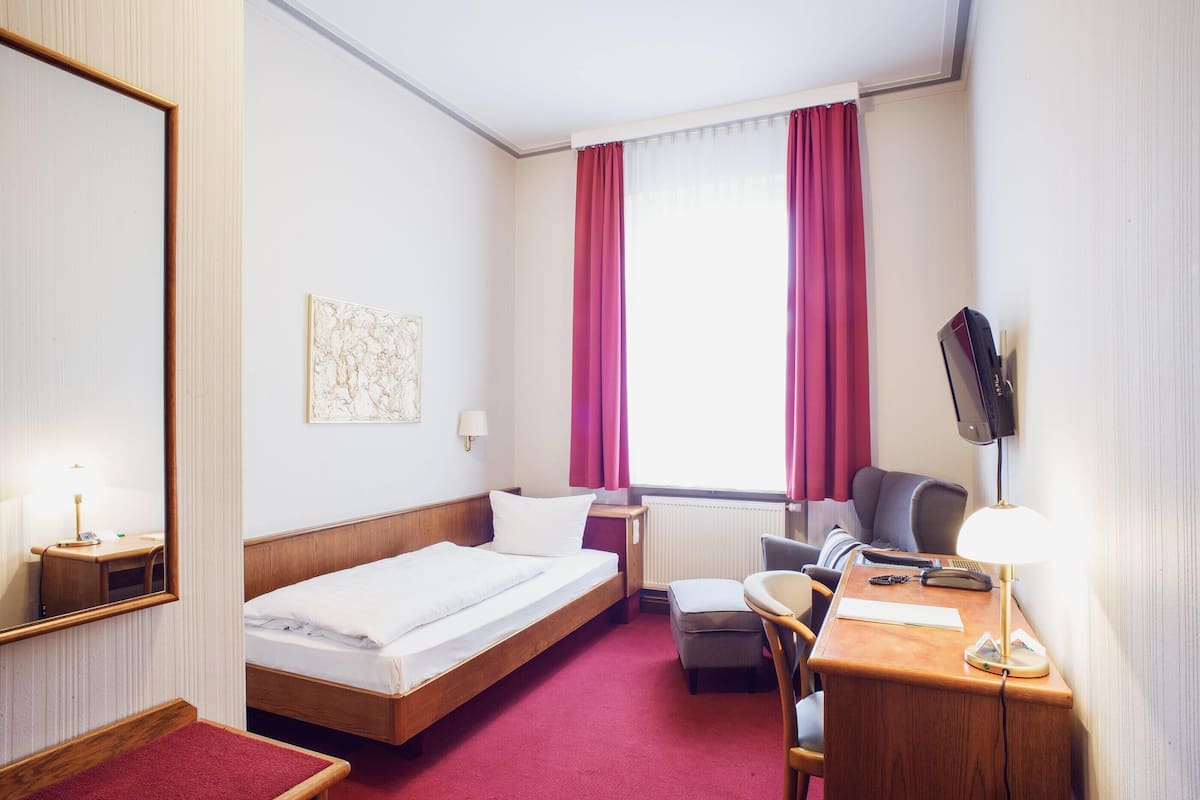 Ein Hotelzimmer mit einem Bett, einem Schreibtisch mit Stuhl, einem an der Wand montierten Fernseher und einem Fenster mit Vorhängen.