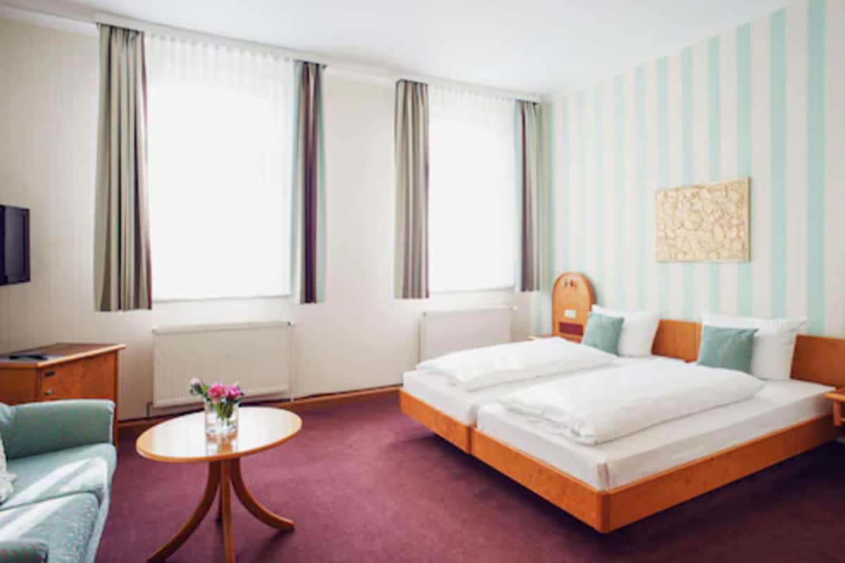 Ein Hotelzimmer mit einem Doppelbett, einem kleinen runden Tisch mit Blumen, einem Nachttisch aus Holz, einem Fernseher und einem Fenster mit Vorhängen.