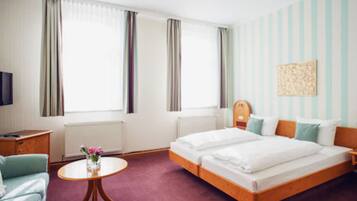 Habitación de hotel con una cama doble, una mesita redonda pequeña con un florero, una mesita de noche de madera, un televisor y una ventana con cortinas.