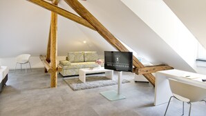 A modern loft-style room with exposed wooden beams, a white desk, a sofa with patterned cushions, and a television displaying 'HOTEL SCHWARZER BAR'. - Hotel Schwarzer Bär (Jena)