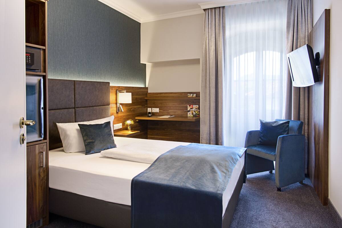 Ein Hotelzimmer mit Bett, Schreibtisch, Stuhl, einem an der Wand montierten Fernseher und einem Fenster mit Vorhängen.