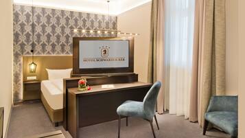 A hotel room with a bed, a desk with a TV displaying the hotel name, a chair, and a sofa.
