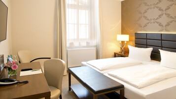 A hotel room with a bed, bedside table, chair, desk, lamp, and a window with curtains.