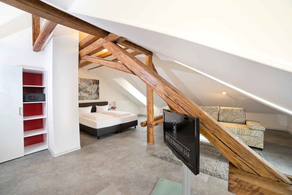 Ein modernes Hotelzimmer mit schrägem Dach, einer Holztreppe, einem Bett, einer Couch und einem roten Stauraum.