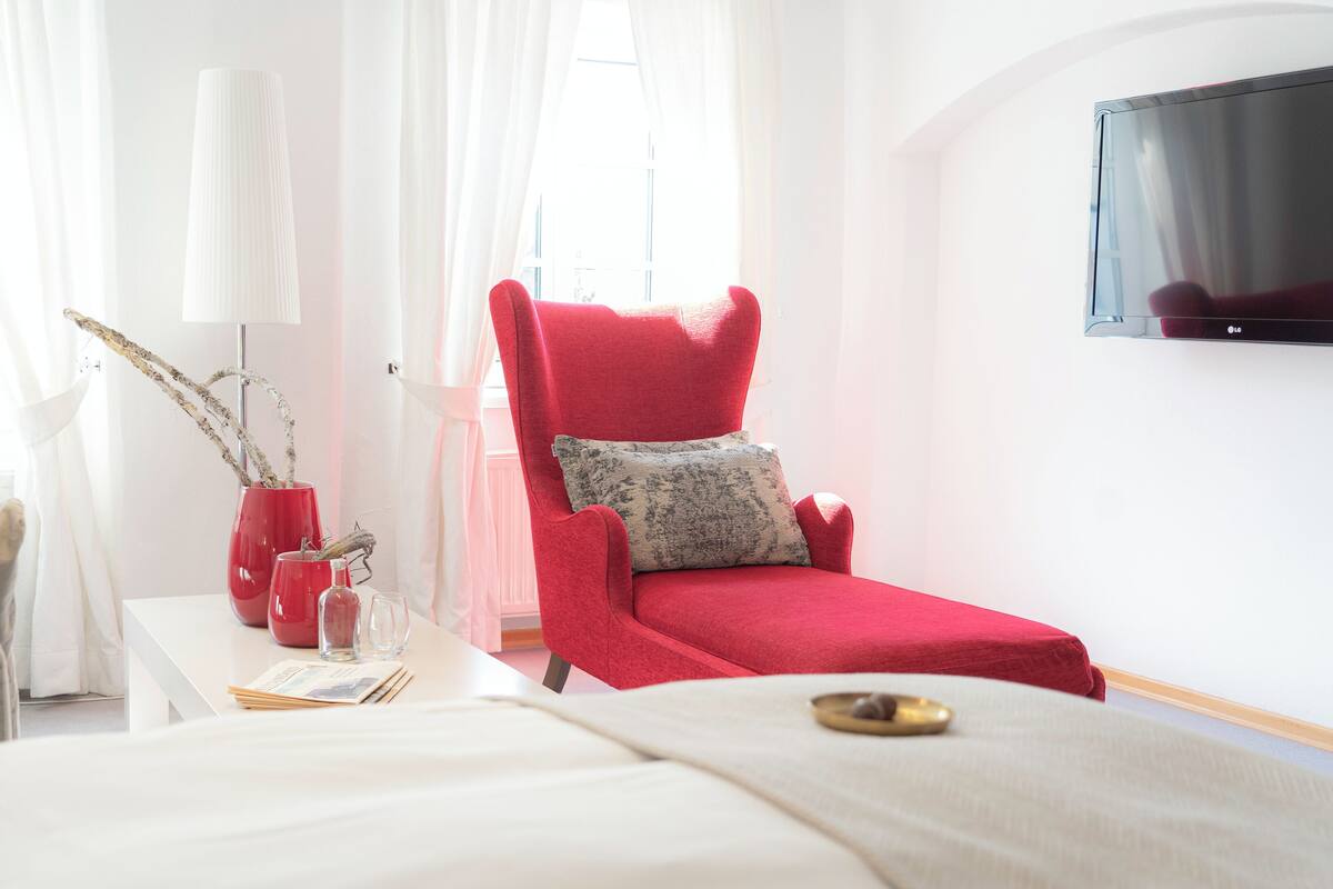 A modern hotel room with a red armchair, a flat-screen TV, a bedside table with a vase and a glass, and a window with curtains.