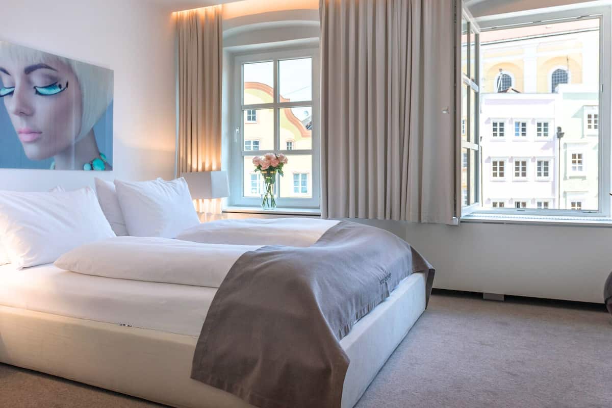 A modern hotel room with a large bed, a bedside table, a window with curtains, and a view of buildings outside.