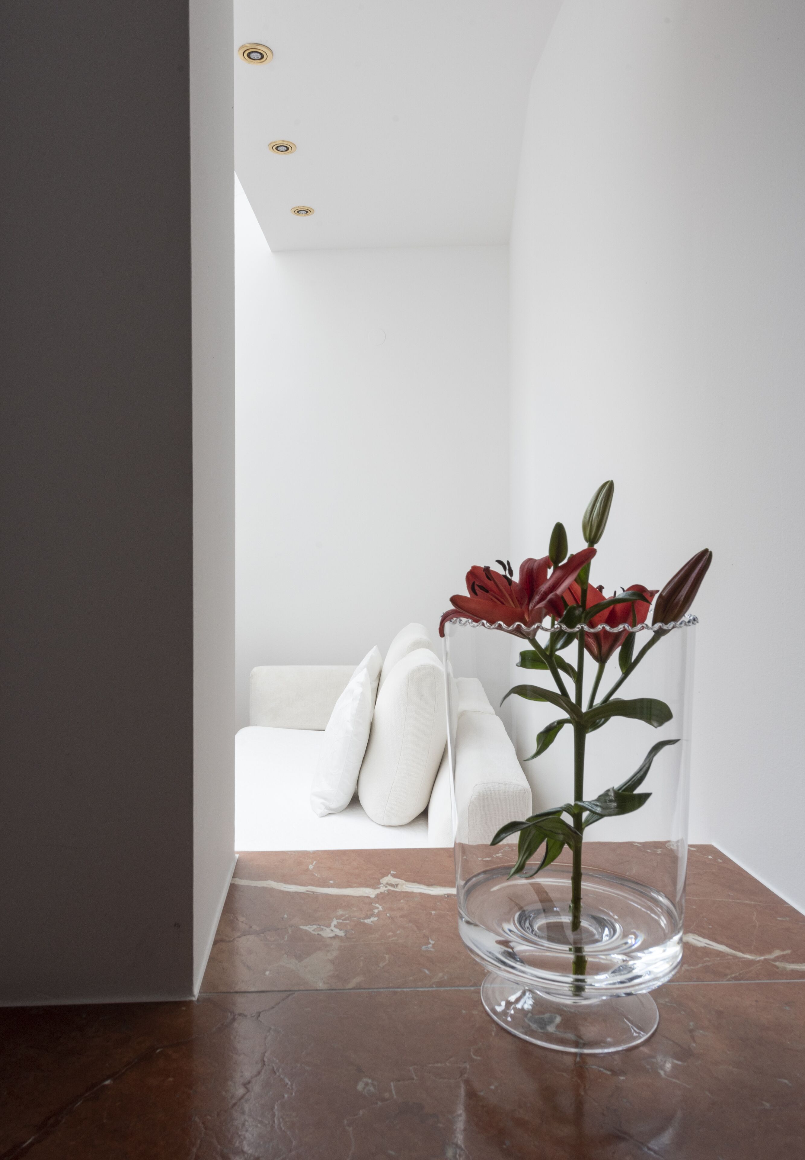 A modern interior with a clear glass vase on a marble floor, a white sofa, and a white wall with recessed lighting.