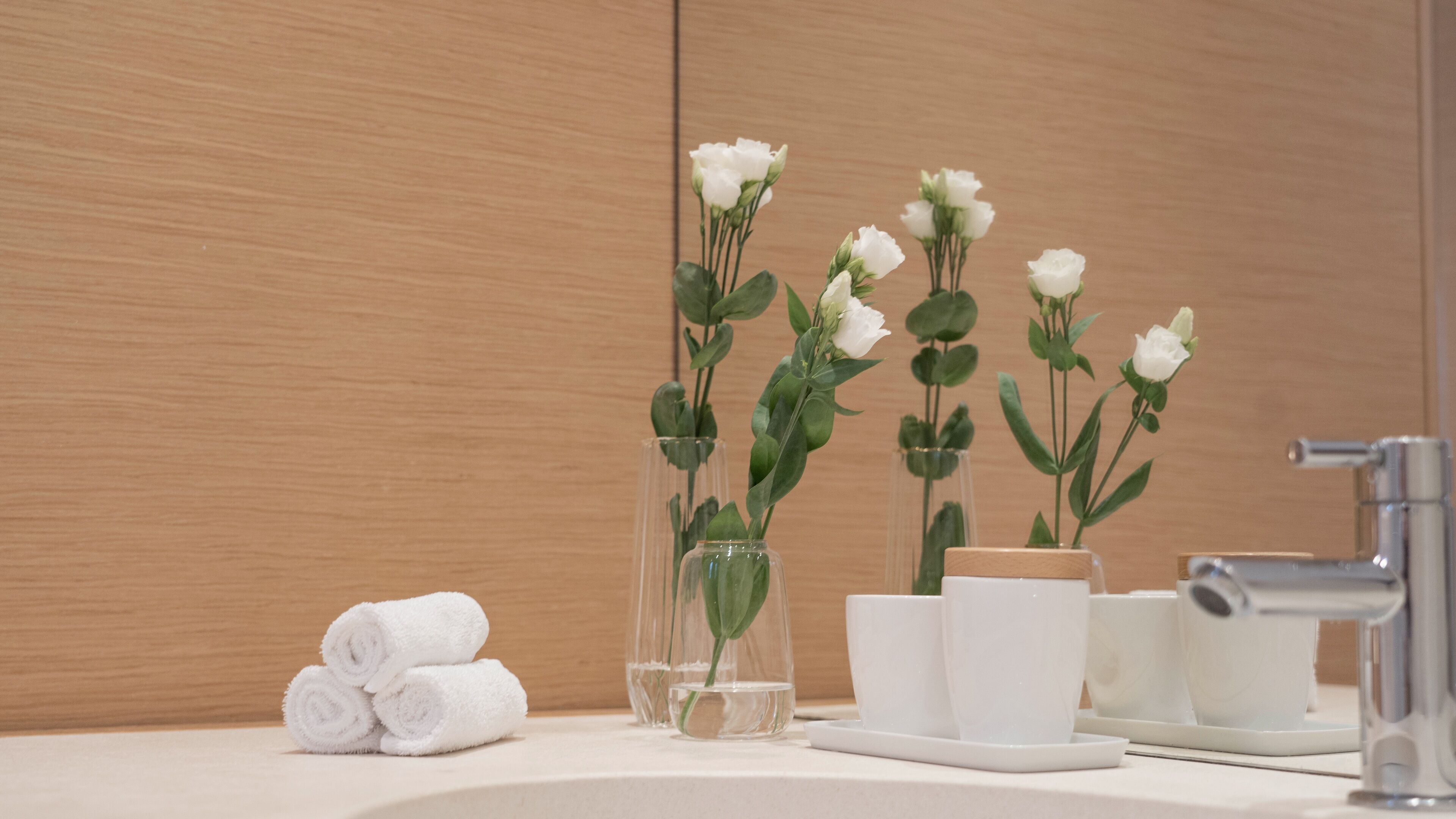 A bathroom with a wooden vanity, white towels, and a vase with white flowers.