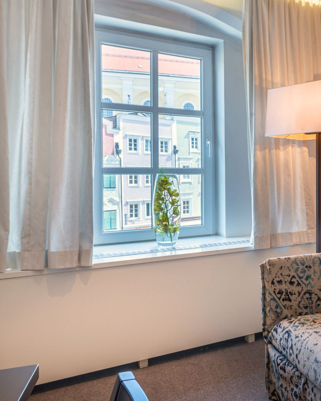 Una habitación con un ventanal amplio, un jarrón con flores y un sillón estampado.