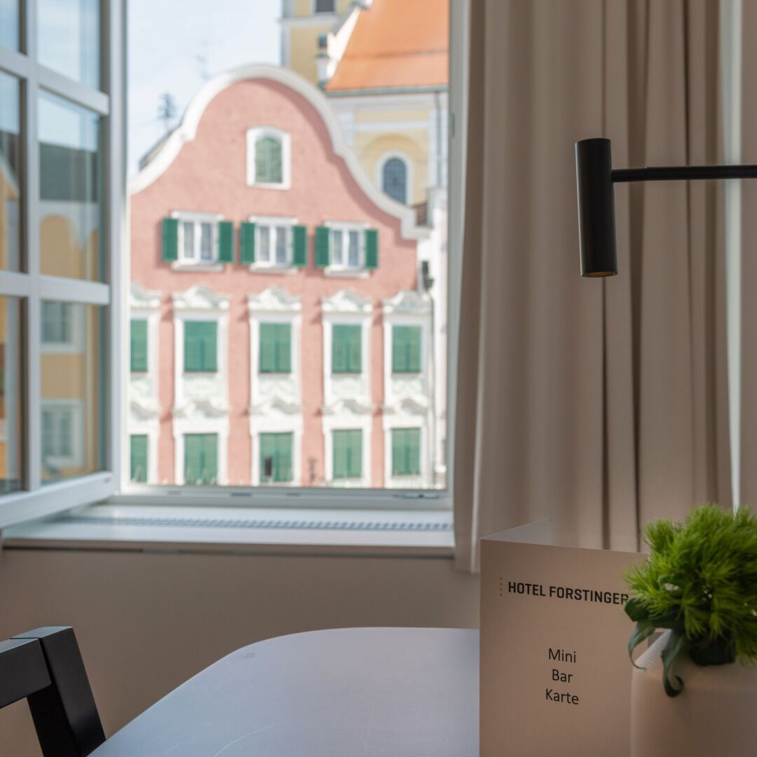 Quarto de hotel com vista para um edifício rosa com persianas verdes, uma planta no parapeito da janela e uma placa indicando 'HOTEL FORSTINGER' com lista de comodidades.