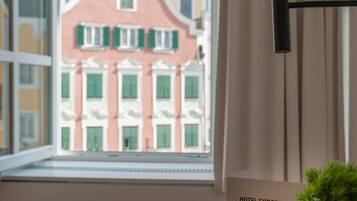 Quarto de hotel com vista para um edifício rosa com persianas verdes, uma planta no parapeito da janela e uma placa indicando 'HOTEL FORSTINGER' com lista de comodidades.