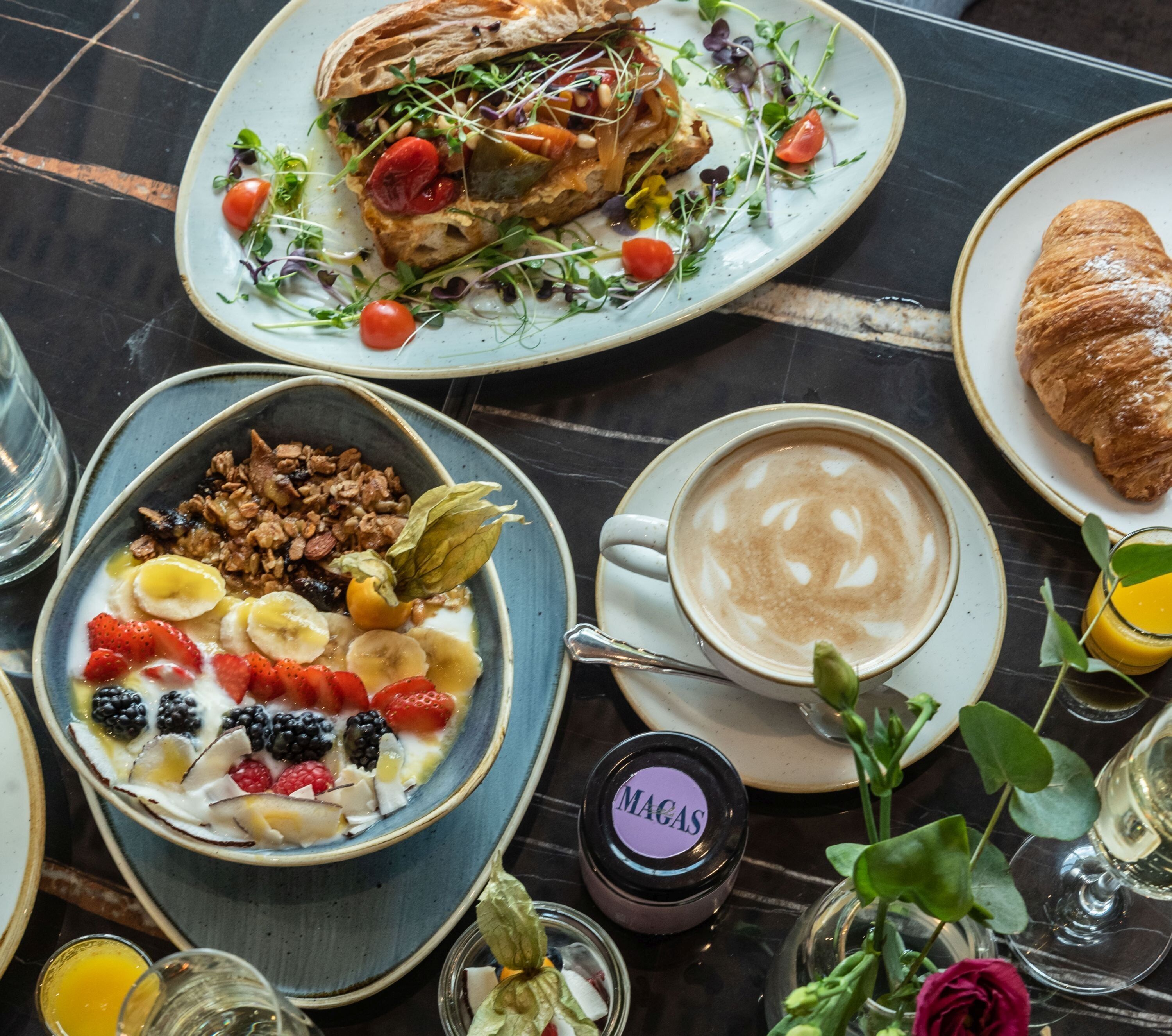 A breakfast setting with a croissant, a bowl of fruit, a cup of coffee, and a plate with a sandwich.