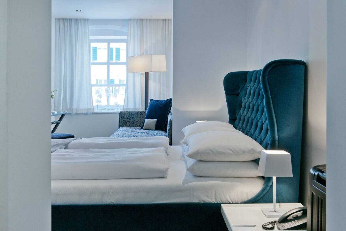 A hotel room with a bed, a desk with a lamp, a telephone, and a window with curtains.