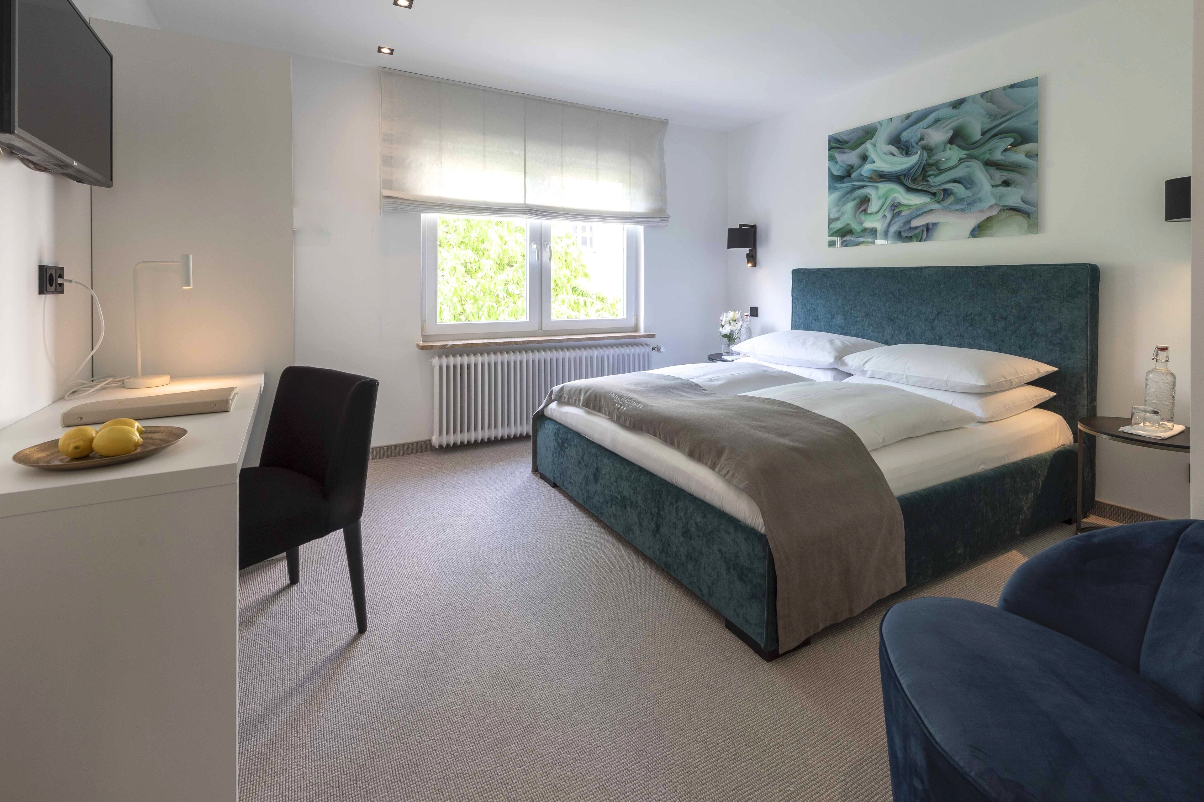 Una habitación de hotel moderna con una cama grande, un escritorio con una silla, una mesita con un florero y una ventana con vista a áreas verdes.