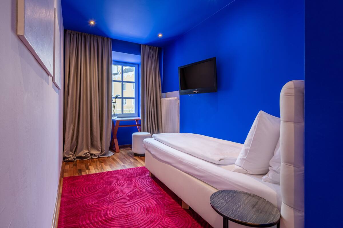 A room with a white sofa, a small table, a TV mounted on the wall, and a window with curtains.