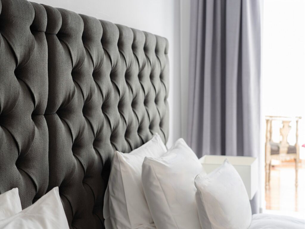 A tufted headboard in a neutral color, white pillows, and a simple bedside table with a lamp.