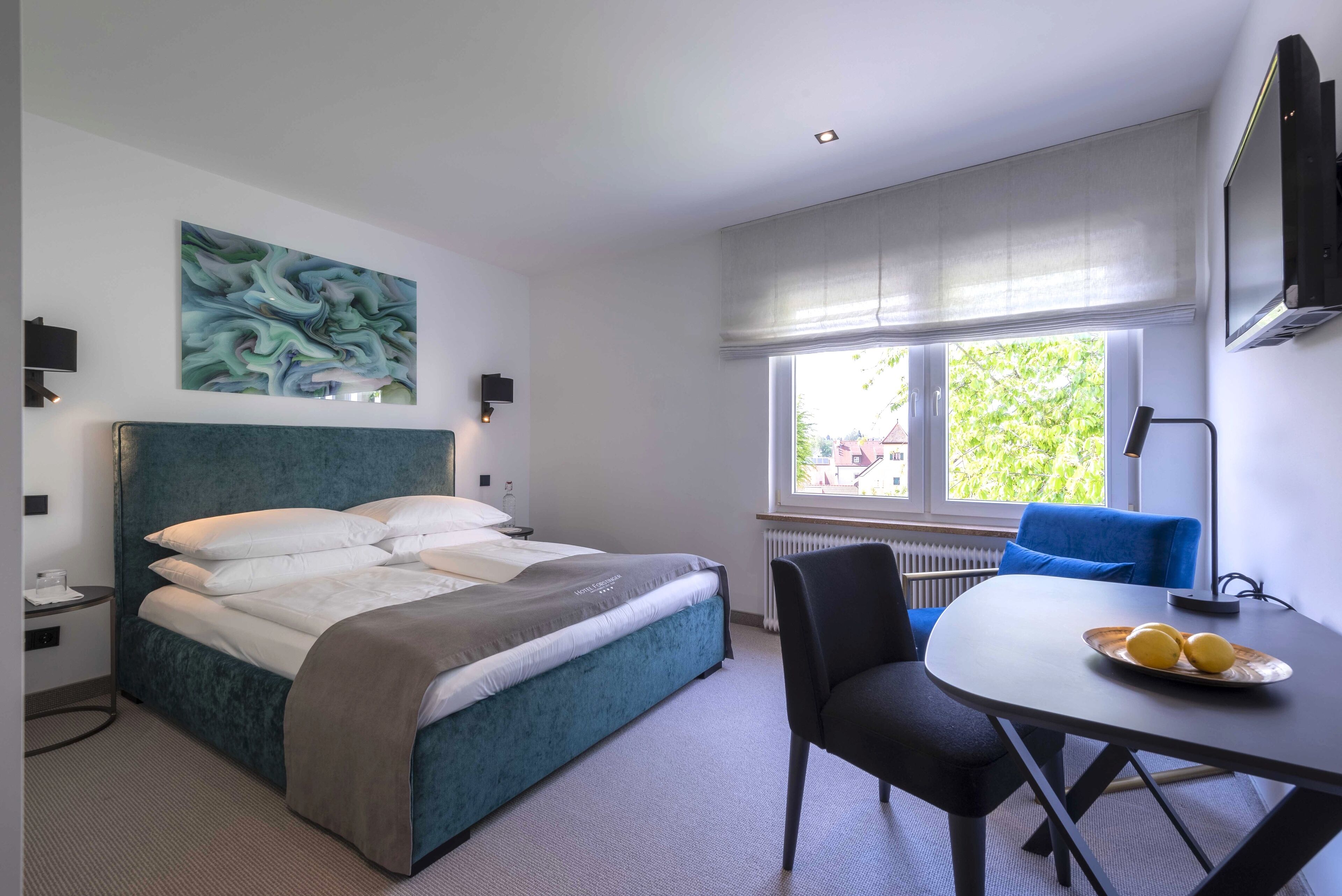 A hotel room with a bed, a table with lemons, a chair, a window with blinds, and a painting on the wall.
