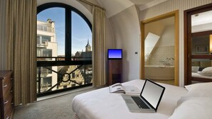 A hotel room with a large arched window, a bed with a laptop, a TV, and a bathroom with a bathtub. - B-aparthotel Ambiorix (Brussels)