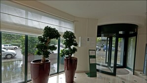 A lobby with two large potted plants, a glass door entrance, and a view of parked cars outside. - B-aparthotel Ambiorix (Brussels)